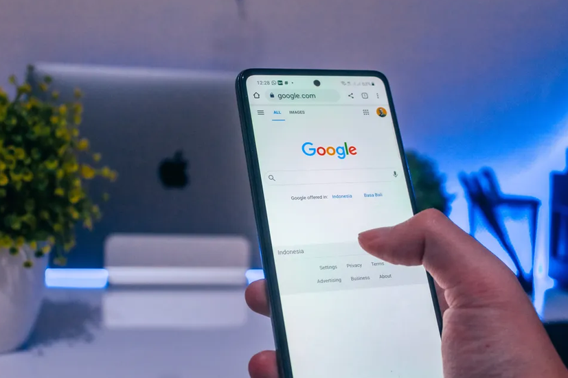 Pessoa segurando um smartphone acessando o site do Google, com um notebook e uma planta desfocados ao fundo em um ambiente iluminado em tons de azul.